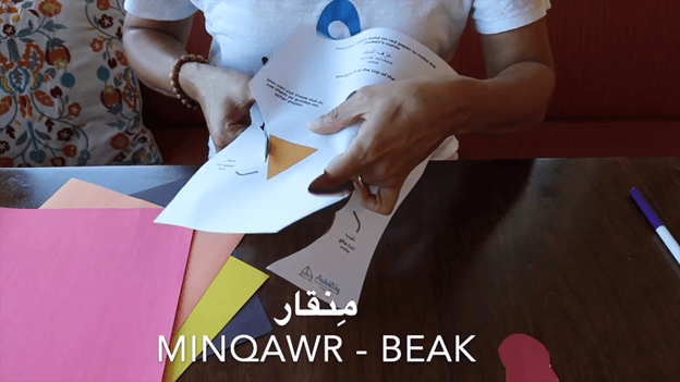 Laila cuts a triangle out of white paper, with Minqawr - Beak in white letters across the bottom 
